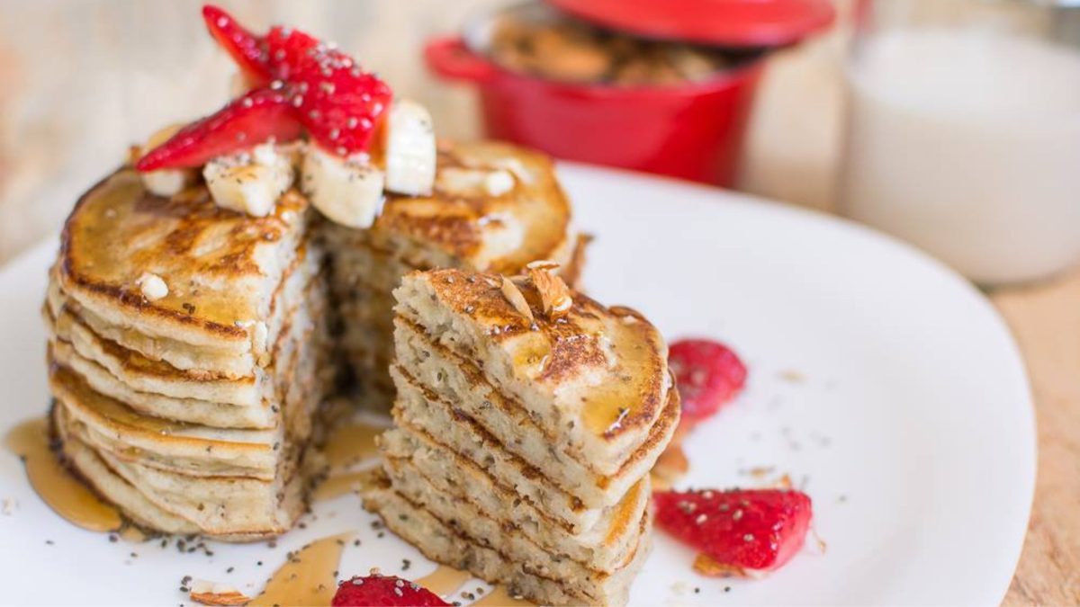 Un plato con una torre de panqueques veganos, cubiertos de syrup y adornados con semillas de chia, trozos de banana y fresa.
